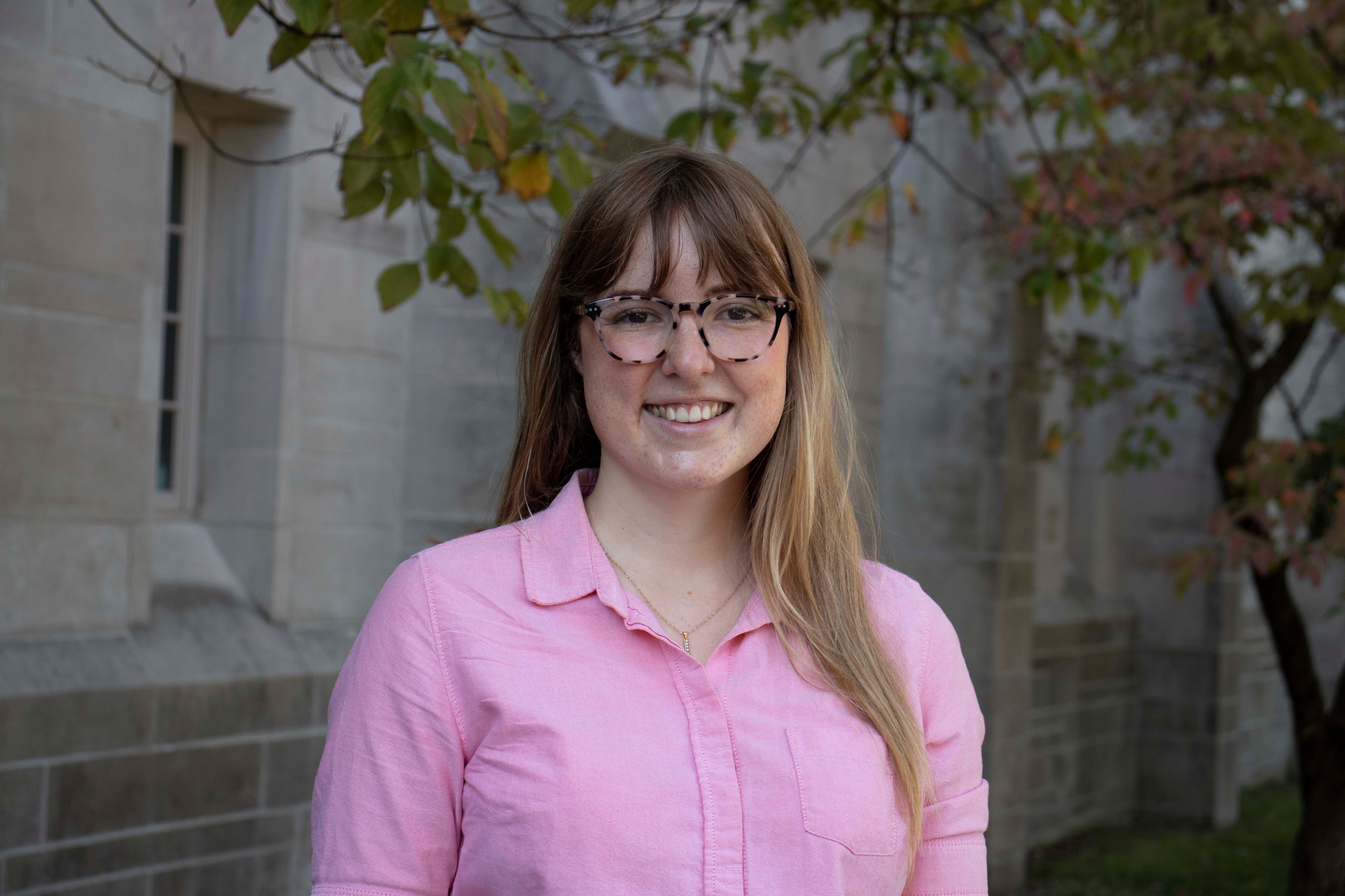A woman wearing a pink shirt and glasses smiles at the camera. Behind her is a limestone building and a tree
